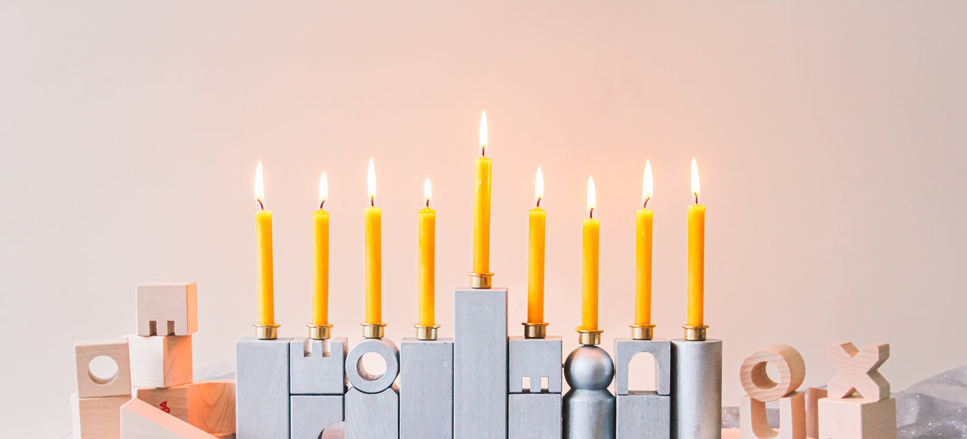 A DIY HABA Wooden Block Menorah sits on a table with lit beeswax candles with unpainted wooden blocks surrounding it.