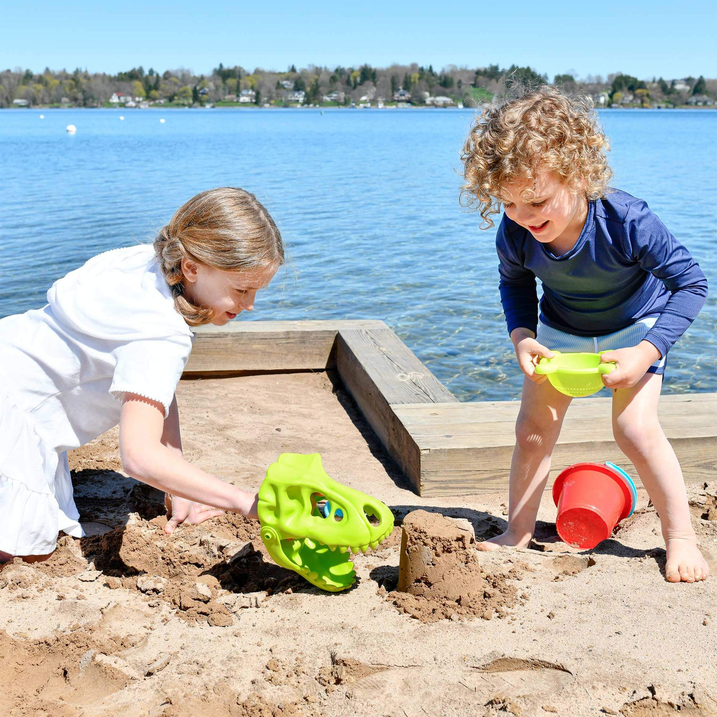 A girl in a white dress playing with green Sand Dino Glove and boy holding green sifter with red bucket laying in the sand and water in the background