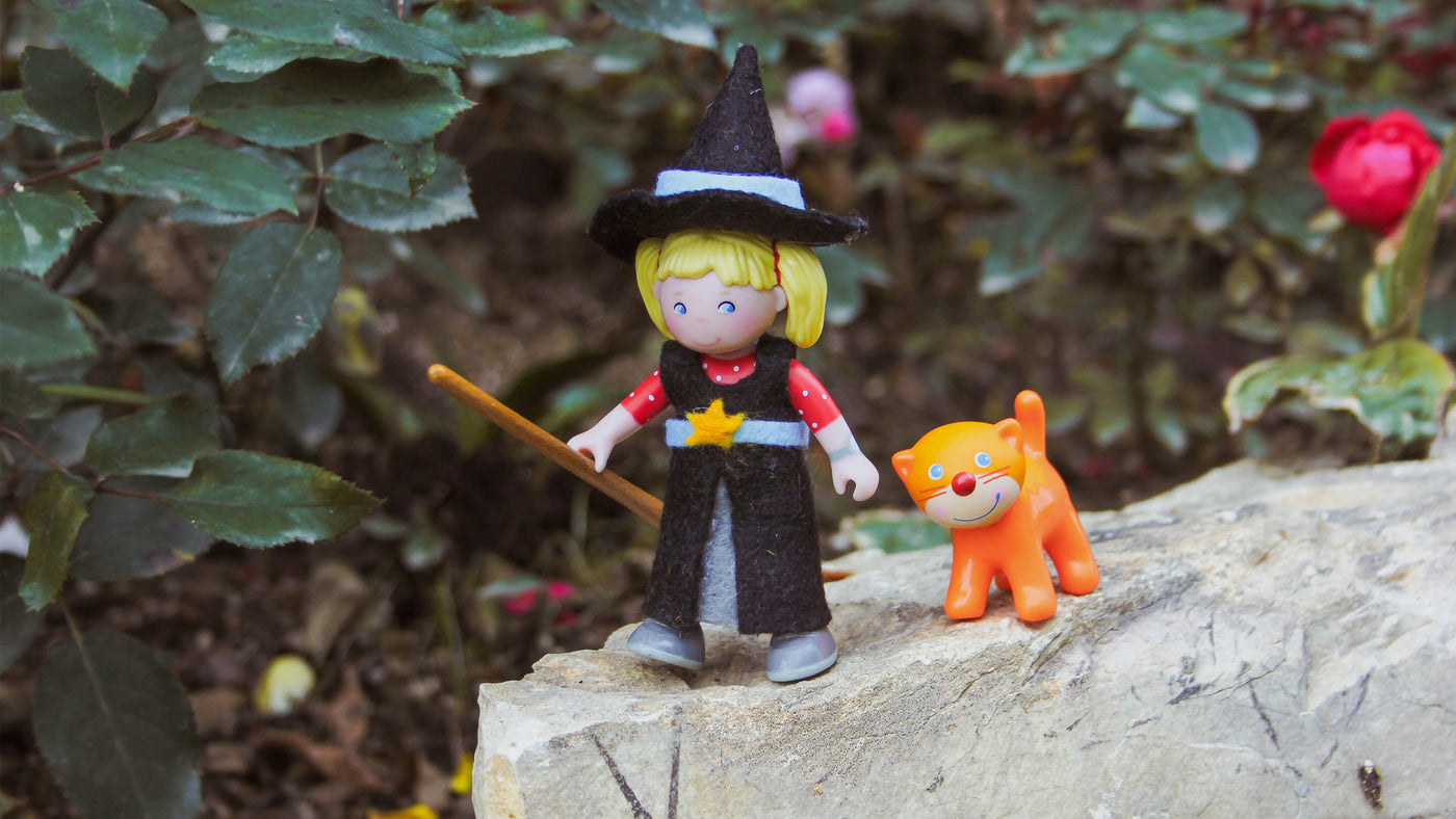 Small vinyl doll dressed in a homemade witch's costume with a little broom and orange cat next to her standing on a stone outdoors