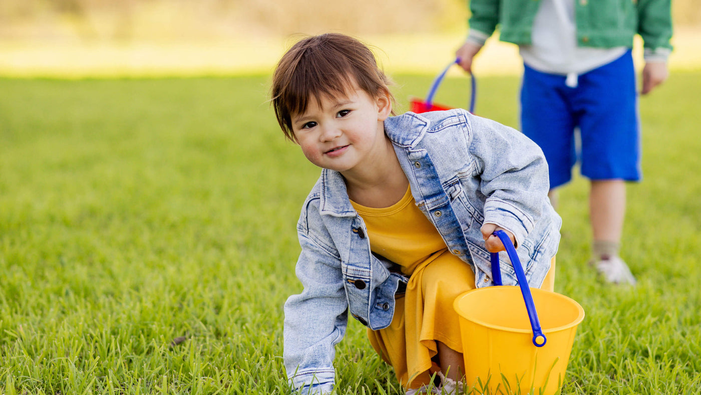 Child in yellow dress with denim jacket holding a yellow bucket on grass.