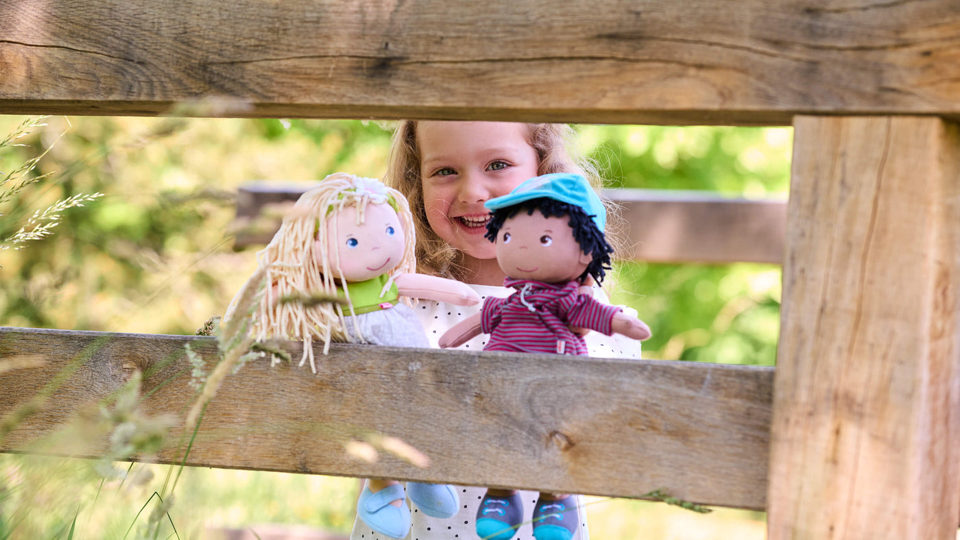 Girl outdoors in summer holding 2 HABA dolls looking at the camera through a fence