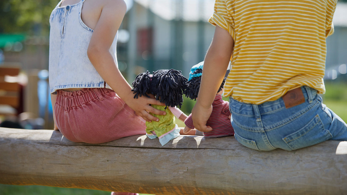 Back view of two kids sitting with HABA dolls outside at a playground