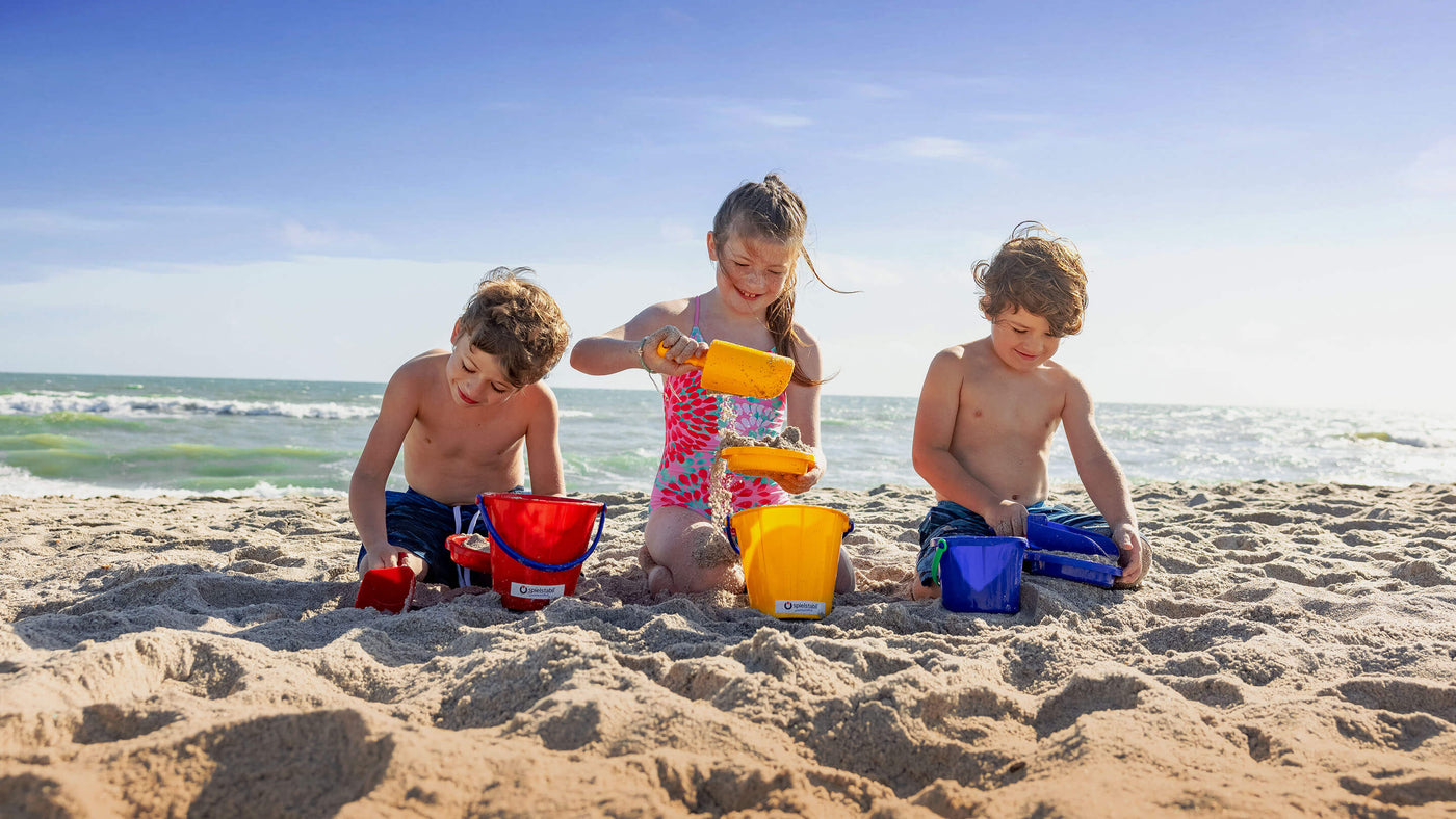 Three kids in the sand at the beach playing with Spielstabil scoops, buckets, and sieves