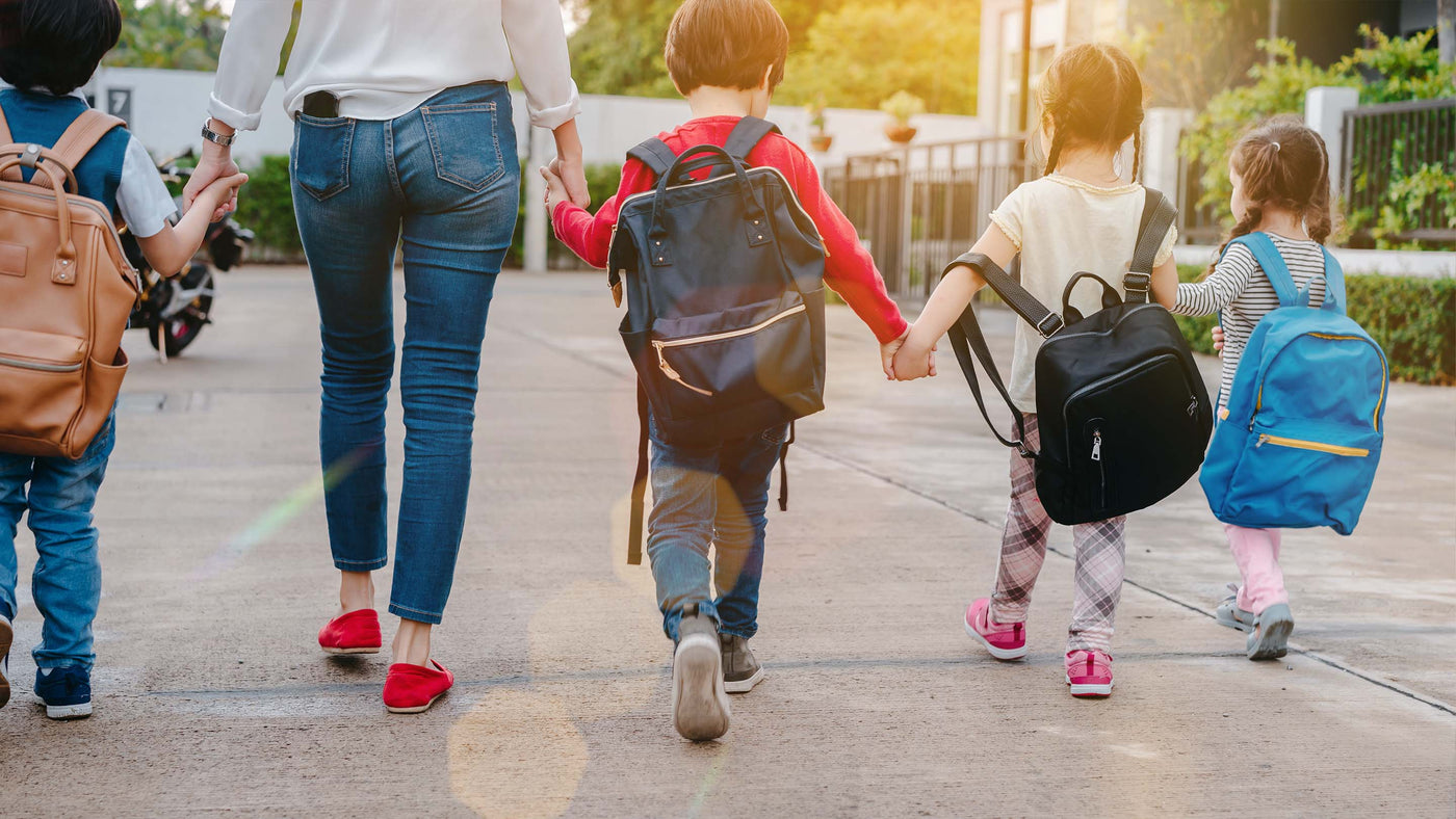 Four kids wearing backpacks holding hands with parent walking away from the camera