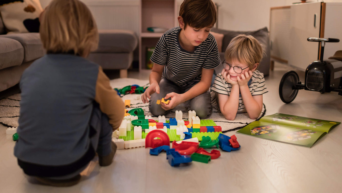 Three children playing with colorful Hubelino building blocks on a hardwood floor.