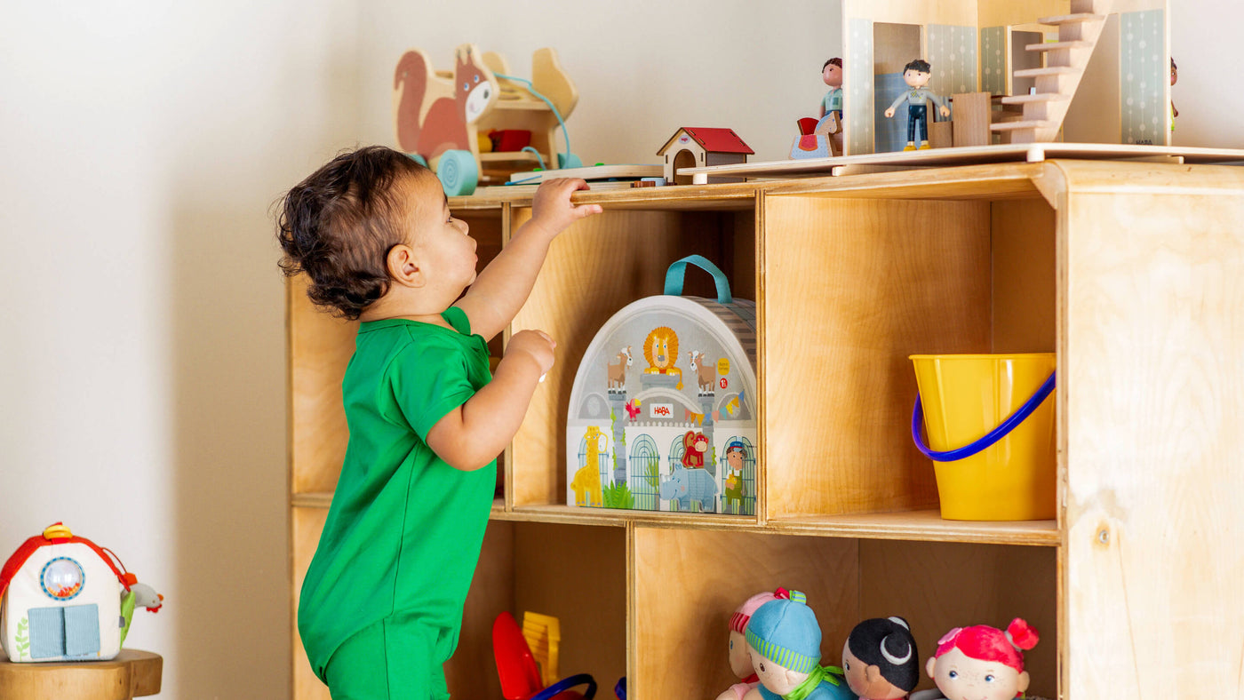 A child in a green onesie reaches for toys on a wooden shelf with various toys and dolls.