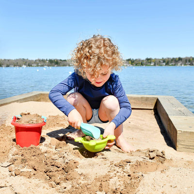 Child playing with Basic Sand Toys Set in the sand with blue shovel and green sifter alongside red bucket filled with sand with water in the background
