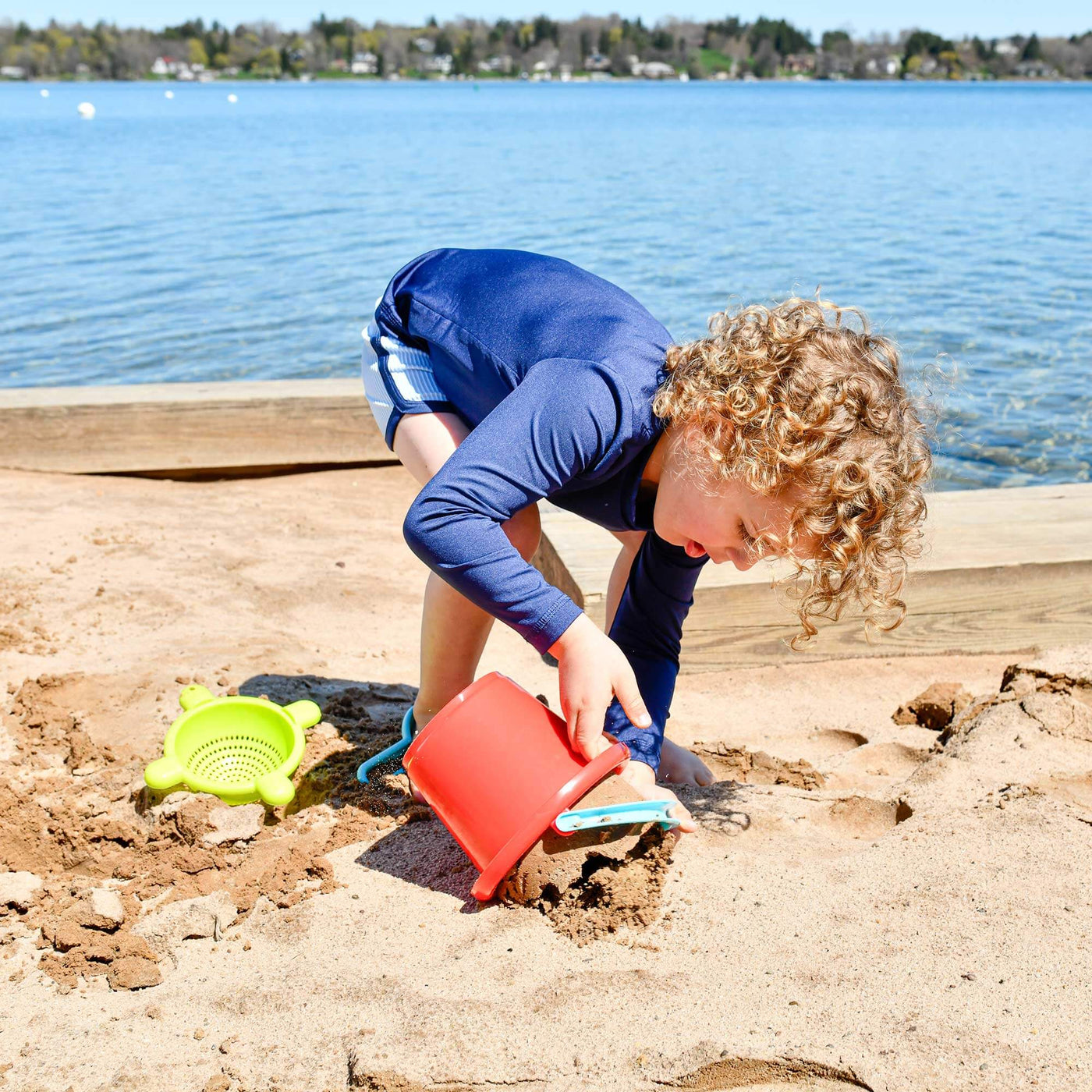 Child playing with Basic Sand Toys Set in the sand with red bucket dumping sand and green sifter with water in the background