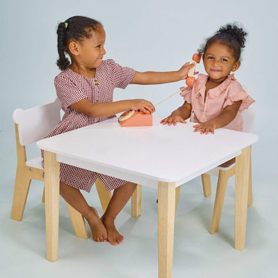 Two young girls playing with a toy at a small wooden table and chairs on a light blue background
