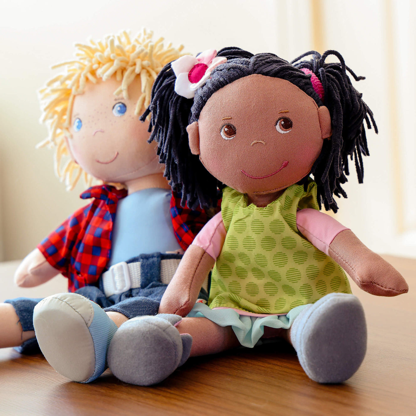 Two fabric dolls sitting on a wooden surface; one has blonde hair and the other has black hair.