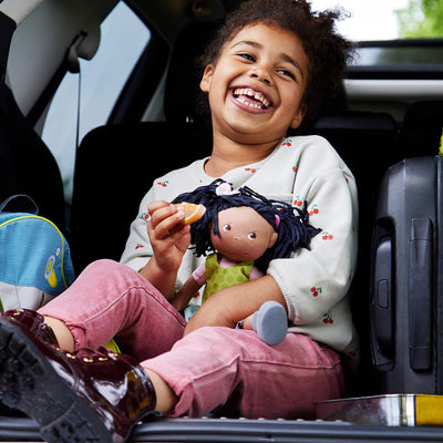 A smiling child sits in a car trunk, holding a Cari doll and wearing a cherry-patterned sweater and pink pants.