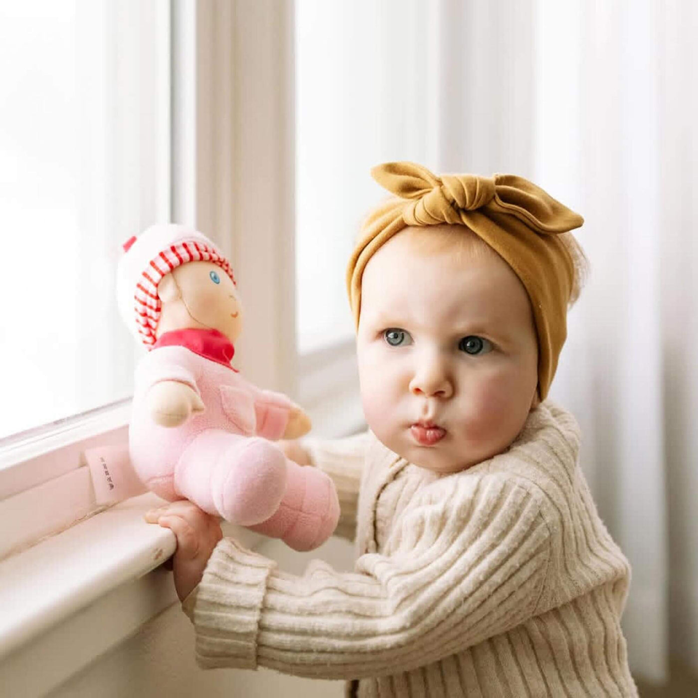 Baby holding a pink doll names Luisa with a bow headband in a softly lit room.