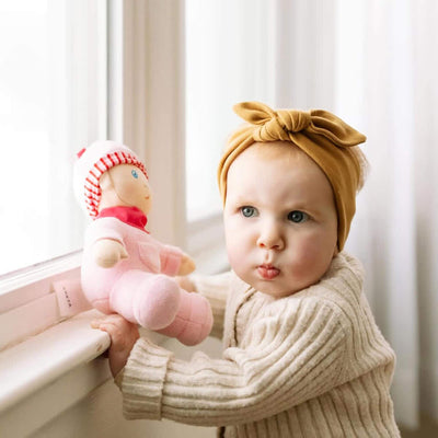 Baby holding a pink doll names Luisa with a bow headband in a softly lit room.