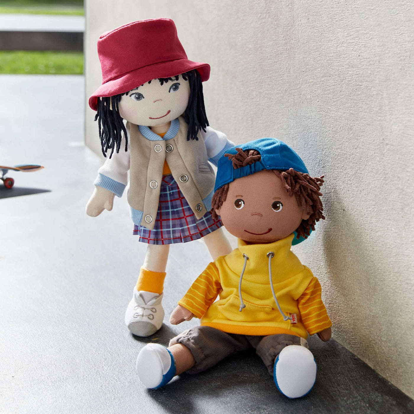 Two dolls posed outside, one standing, wearing a red hat and plaid skirt, the other sitting with a blue cap and yellow hoodie.