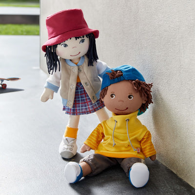 Two dolls posed outside, one standing, wearing a red hat and plaid skirt, the other sitting with a blue cap and yellow hoodie.