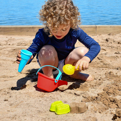 A child with curly hair plays in the sand, holding a blue cone and yellow shovel next to a red bucket and colorful molds.
