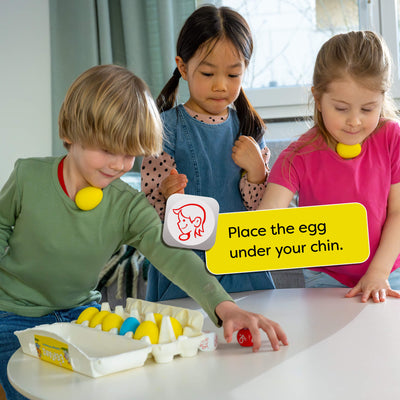 Three children playing with HABA Dancing Eggs at a table, with a sign instructing to 'Place the egg under your chin'.