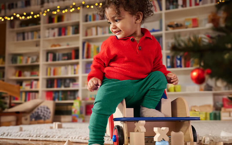 Toddler standing on HABA Walker Wagon Push Toy surrounded by wooden blocks in front of a Christmas tree