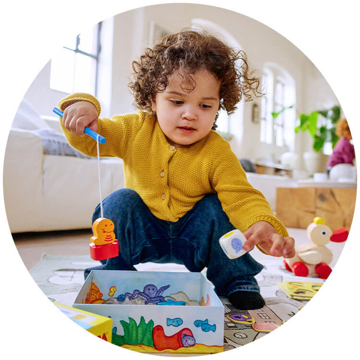 A cheerful toddler in a yellow sweater plays with My Very First Games Here Fishy, Fishy! fishing game, catching colorful characters from a box on a play mat.