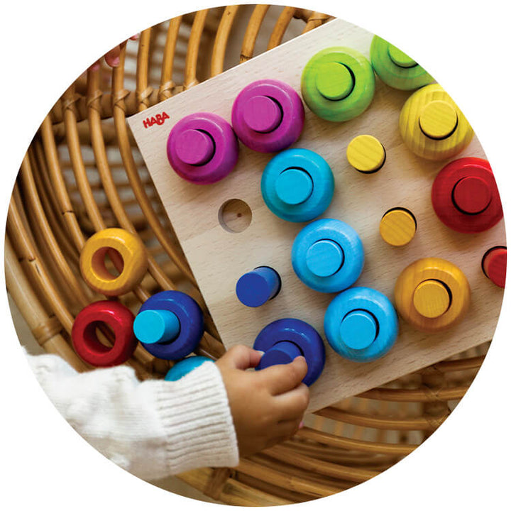A child's hand reaches for colorful wooden pegs on Rainbow Whirls Wooden Sorting and Stacking Game, set against a textured rattan background.