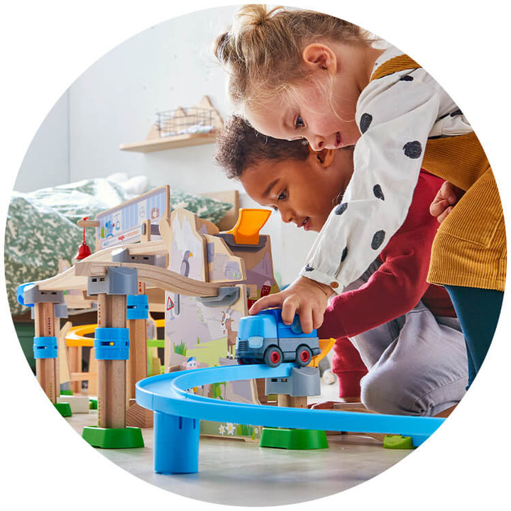 Two kids play together with a Kullerbu Mountain Adventure Track Set, carefully guiding a blue train along a curved track in a bright room.