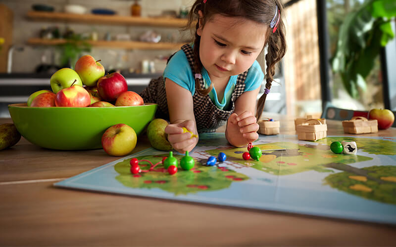 Child playing with Orchard board game on a kitchen counter with apples and a green bowl.