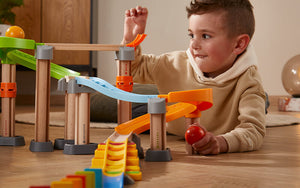 A young boy plays with a Kullerbu Melody Dominoes Set, smiling as he adjusts the tracks and holds a red ball in a cozy room.