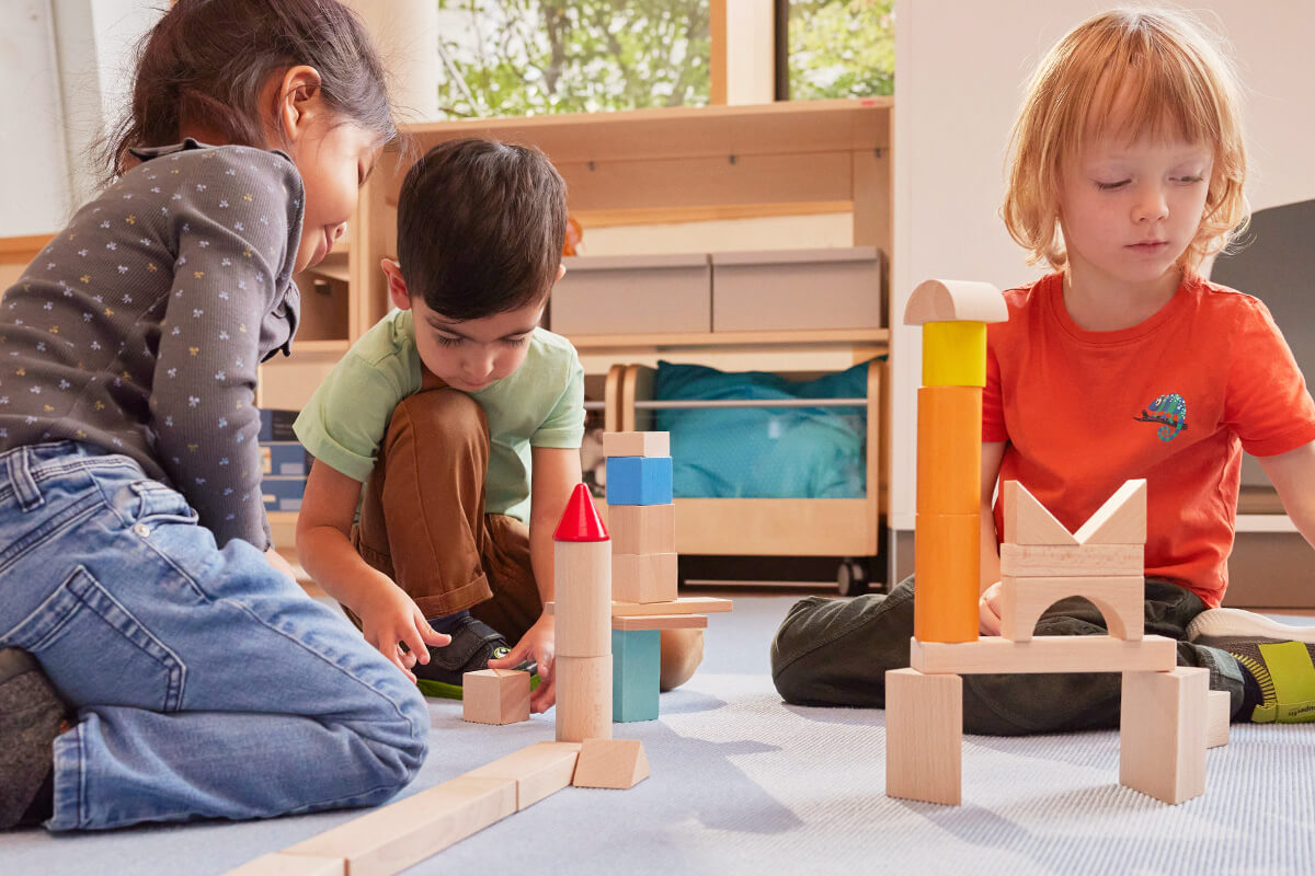 Three children playing with HABA wooden blocks on a carpeted floor in a sunlit library room.