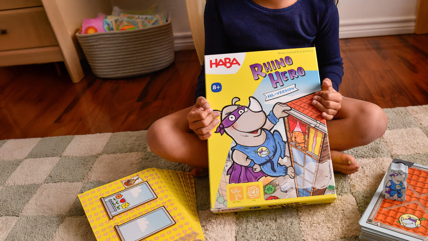 Kid sitting on a rug holding an oversized game box called HABA Giant Rhino Hero XXL