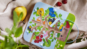Close up of Haba's Orchard Magnetic Maze Game on fabric in a basket with cherries and a pear