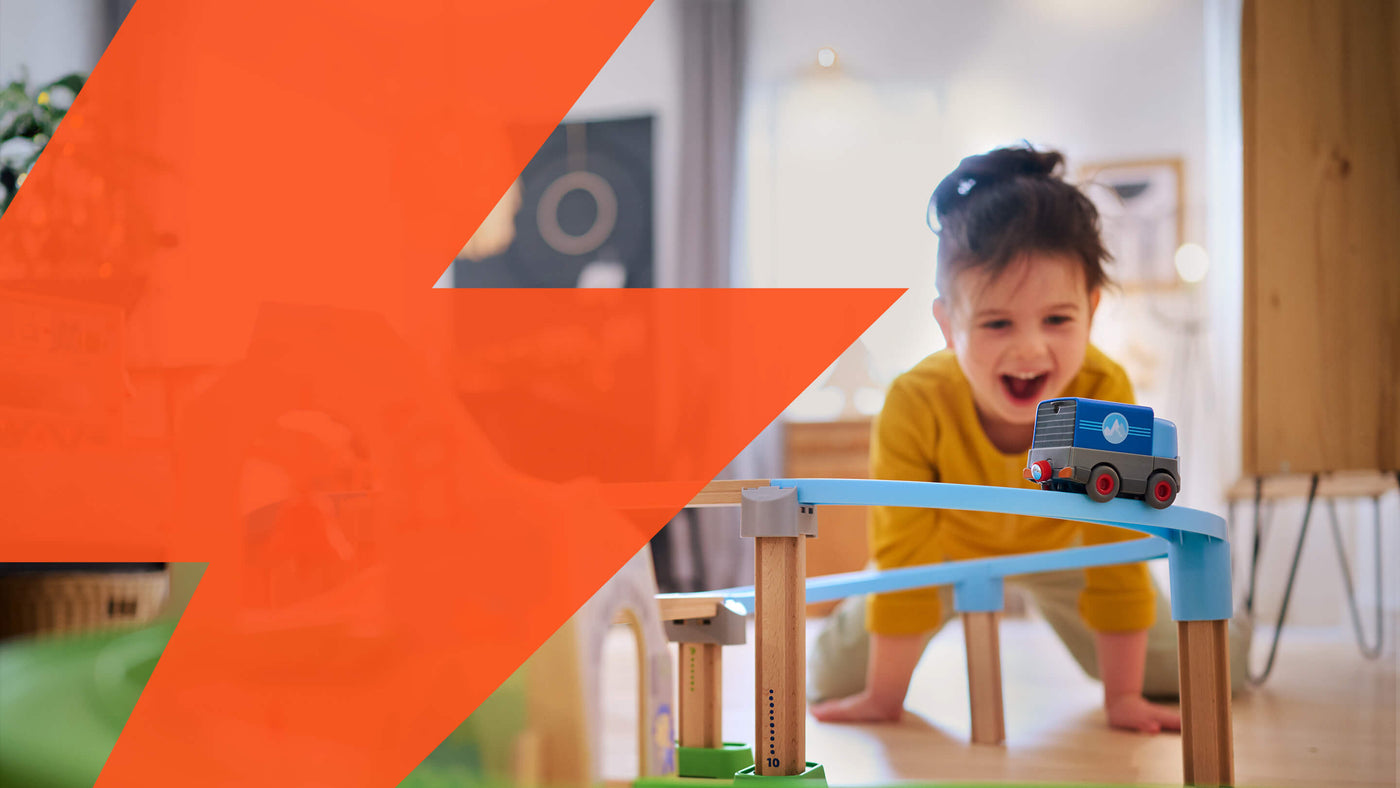 A child in a yellow top watches a toy truck from the HABA Kullerbu Mountain Adventure Track Set in a room, with an orange geometric shape overlaying the left side of the image.