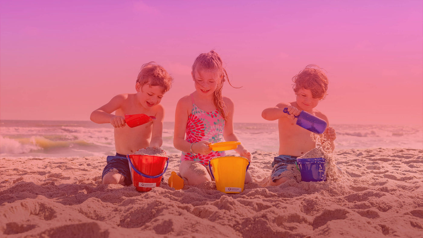 Three children playing with sand buckets and shovels on a beach at sunset.