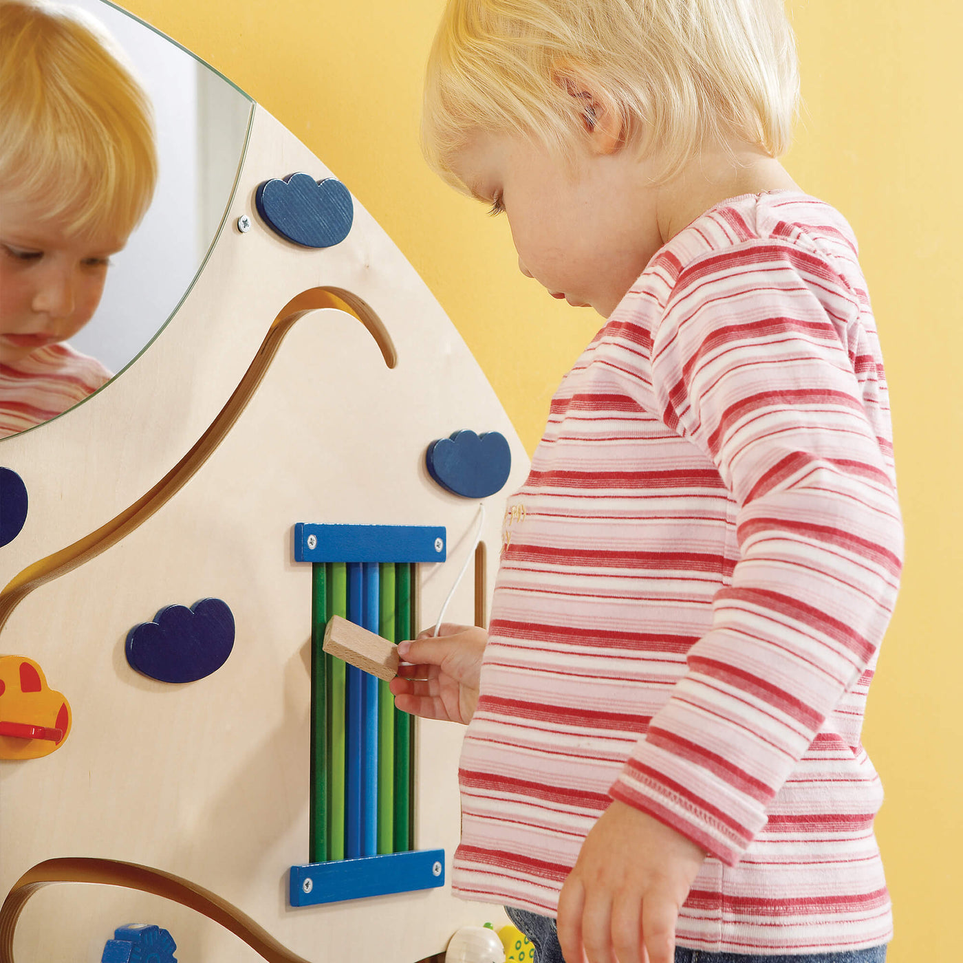 A child holding a tethered small wooden block while looking closely at a section of the HABA Sensory Learning Wall Panel  that includes vertical green and blue rods.