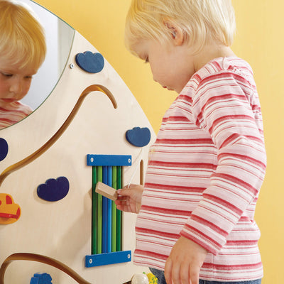 A child holding a tethered small wooden block while looking closely at a section of the HABA Sensory Learning Wall Panel  that includes vertical green and blue rods.