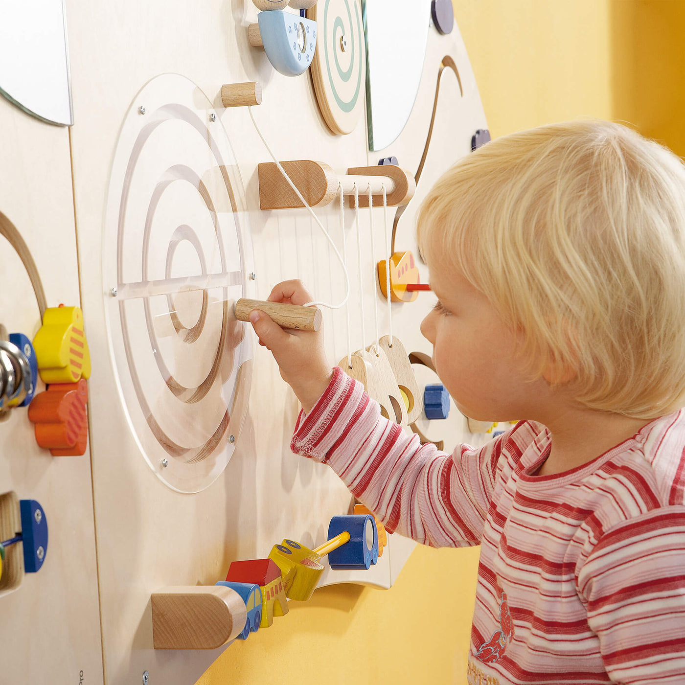 Wood Sensory Learning Wall Panel featuring a circular puzzle with concentric rings covered by transparent plastic, which the child is engaging with a wooden stylus.
