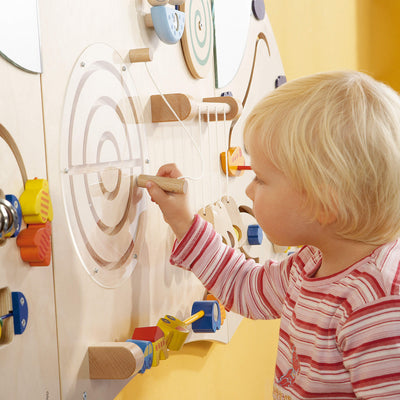 Wood Sensory Learning Wall Panel featuring a circular puzzle with concentric rings covered by transparent plastic, which the child is engaging with a wooden stylus.