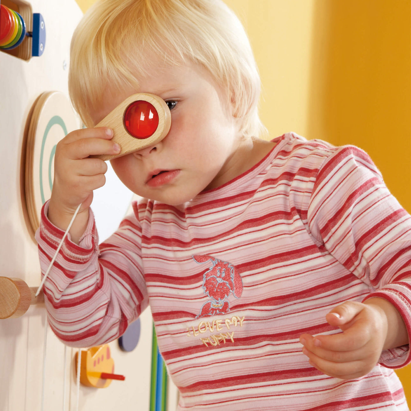 Child looking through a wooden toy with a red lens that is tethered to the HABA Sensory Learning Wall Panel 