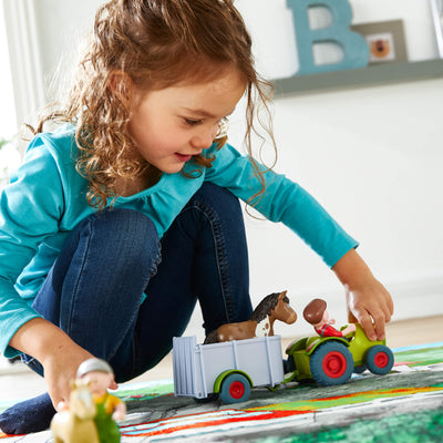 Girl crouching down to push a Little Friends tractor with a toy farmer figure in it attached to the trailer with a toy horse in it across a carpet