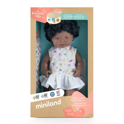 Miniland Montessori Black girl doll with curly hair and yellow bow, wearing a floral top and white skirt, standing in the box she is packaged in. 