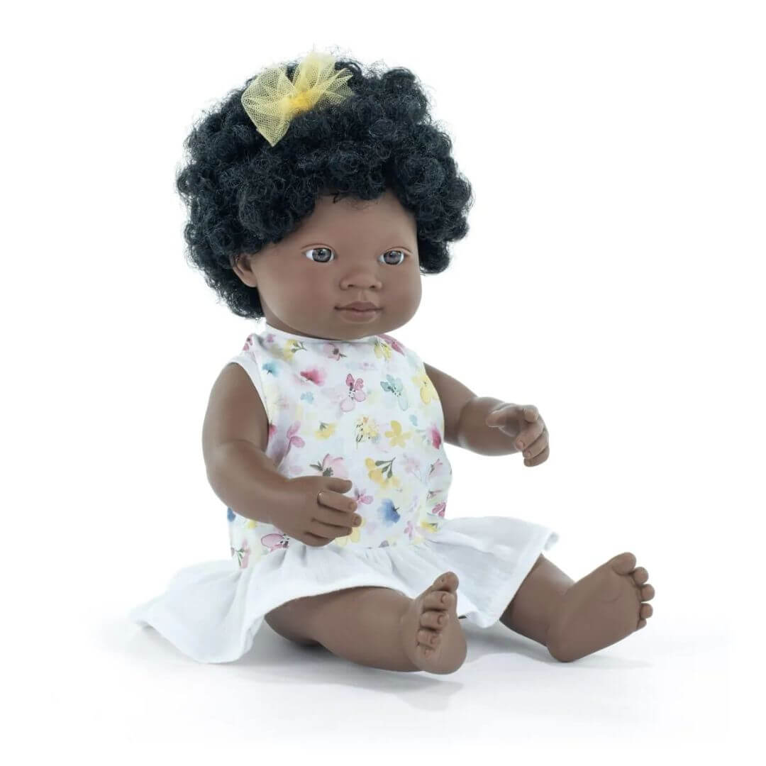 Miniland Montessori Black girl doll with curly hair and yellow bow, wearing a floral top and white skirt, seated on a white background.