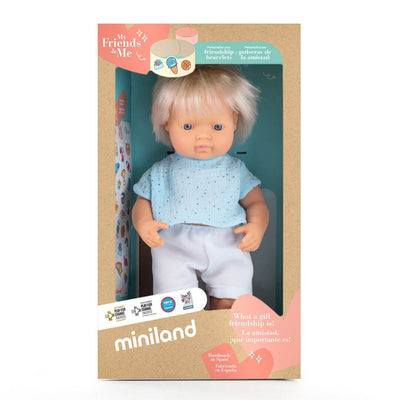 A realistic Miniland Montessori white boy doll with blond hair, wearing a light blue short-sleeved outfit, standing in the packing he arrives in. 