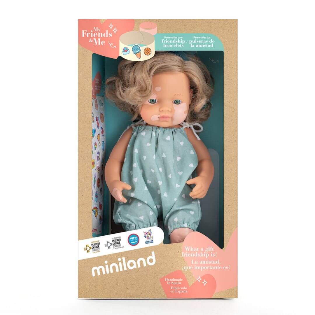 Miniland Realistic Montessori Doll of a white girl with blonde curly hair and vitiligo, wearing a teal polka-dot romper and matching bow, standing in the box she comes packaged in.