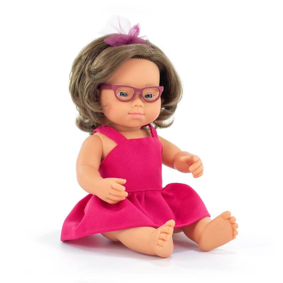 iniland realistic Montessori girl doll with Down syndrome, wearing glasses and a bright pink dress with a tulle bow headband, seated against a white background.