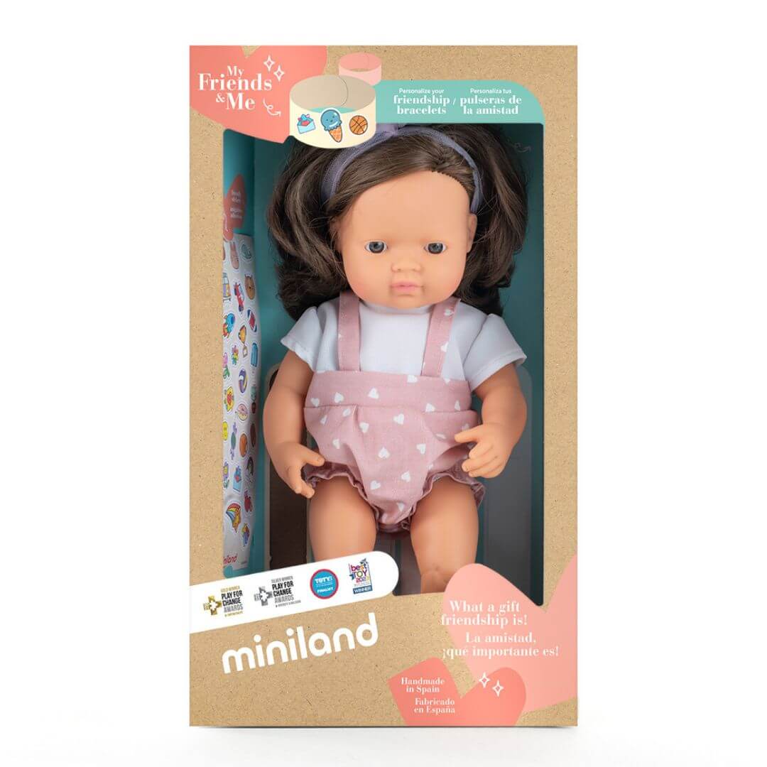 Miniland Montessori girl doll with soft brown hair, wearing a pink polka dot romper and headband, standing in the box she is packaged in.