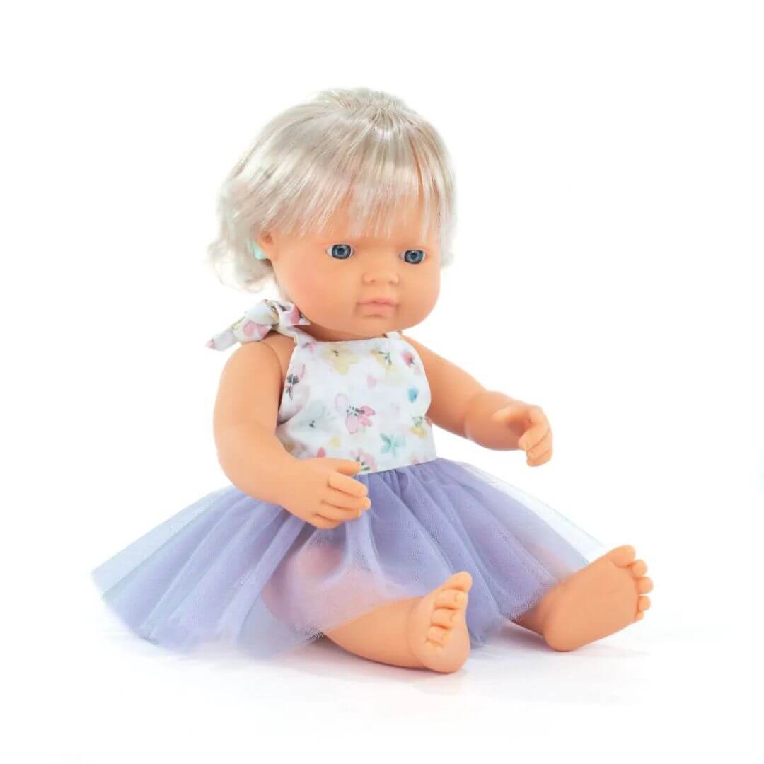 Miniland Montessori doll with blonde hair and visible cochlear implant, wearing a floral top and lavender tulle skirt, seated on a white background.