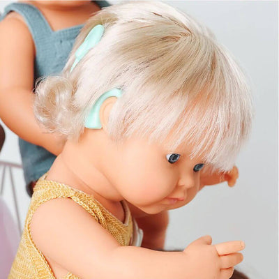 A close up of the cochlear implant on this Miniland Montessori doll with blonde hair wearing a yellow knit top and another doll in the background.