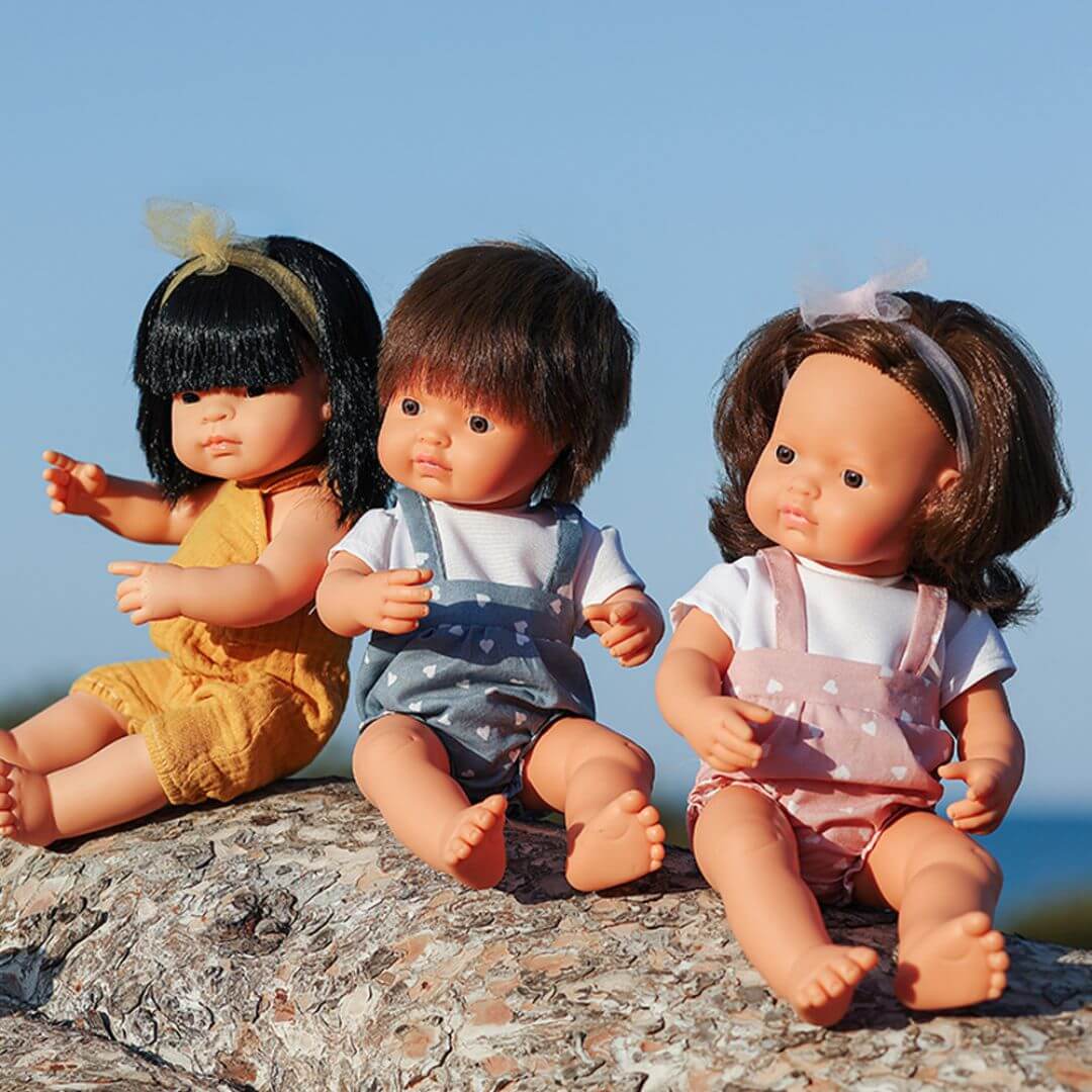 Three Miniland Montessori dolls sitting on a rock outdoors against a blue sky backdrop. The dolls represent diverse ethnicities and wear colorful summer outfits, each posed with expressive hand gestures and realistic features.