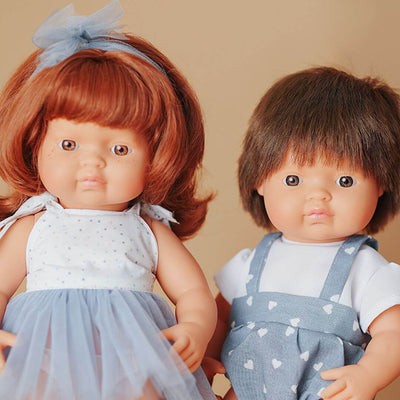 Miniland Montessori red-haired girl doll in a white top and blue skirt standing next to a boy doll in a white shirt and blue overalls with heart pattern.
