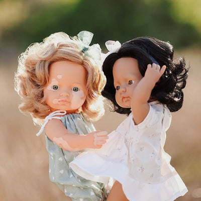 Two Miniland realistic Montessori dolls, one with vitiligo and curly blonde hair wearing a green dress, and one Hispanic doll with dark hair in a white dress, standing together outdoors.