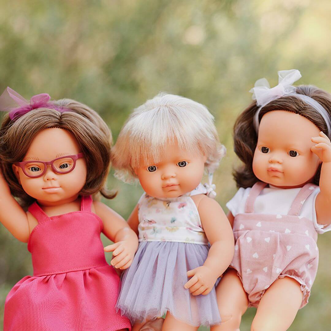 Three Miniland Montessori dolls standing outdoors with blurred trees as a backdrop. The dolls wear colorful summer outfits, each posed with expressive hand gestures and realistic features.